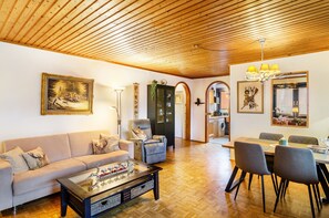 Un salon comprenant un canapé, un fauteuil, une table basse, une table à manger avec des chaises, un plafond en bois et, au fond, une cuisine.