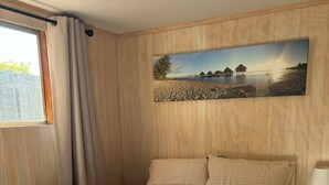 A bedroom with a bed, a painting of a beach scene, a window with curtains, and a bedside table with a lamp.