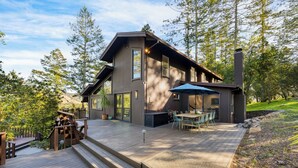 A modern two-story house with a deck, outdoor seating, and a large window overlooking a forested area. - 10-Acre Wellness Retreat, Pool, Sauna, Ceremony Tent | Fable Sonoma by AvantStay (Santa Rosa)