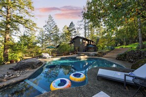 Une piscine dotée de chaises longues et de bouées gonflables, entourée d’arbres et d’un chalet en bois.