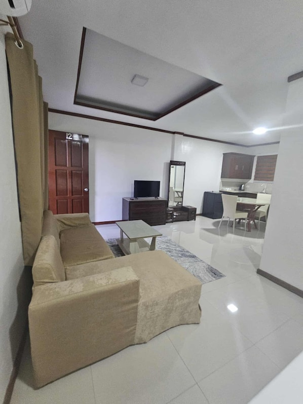 A modern living room with a beige sectional sofa, a flat-screen TV, a dining table with chairs, and a kitchen area with cabinets and a countertop. - Bliss Suites by SMS Hospitality (Panglao)
