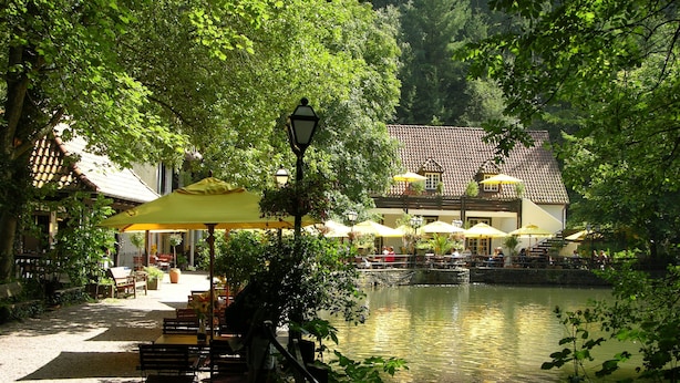 A riverside restaurant with outdoor seating, a bridge, and a building with a tiled roof in the background. - Waldhotel Silbermühle (Horn-Bad Meinberg)