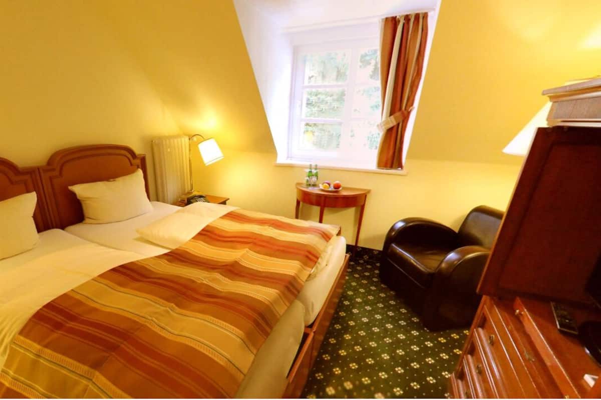 Een hotelkamer met een bed, een stoel, een tafeltje, een lamp en een raam met gordijnen.