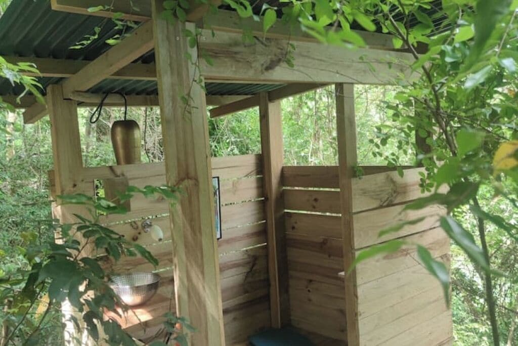 A wooden structure with a metal container hanging from the ceiling, a metal bowl on the floor, and a bench inside.