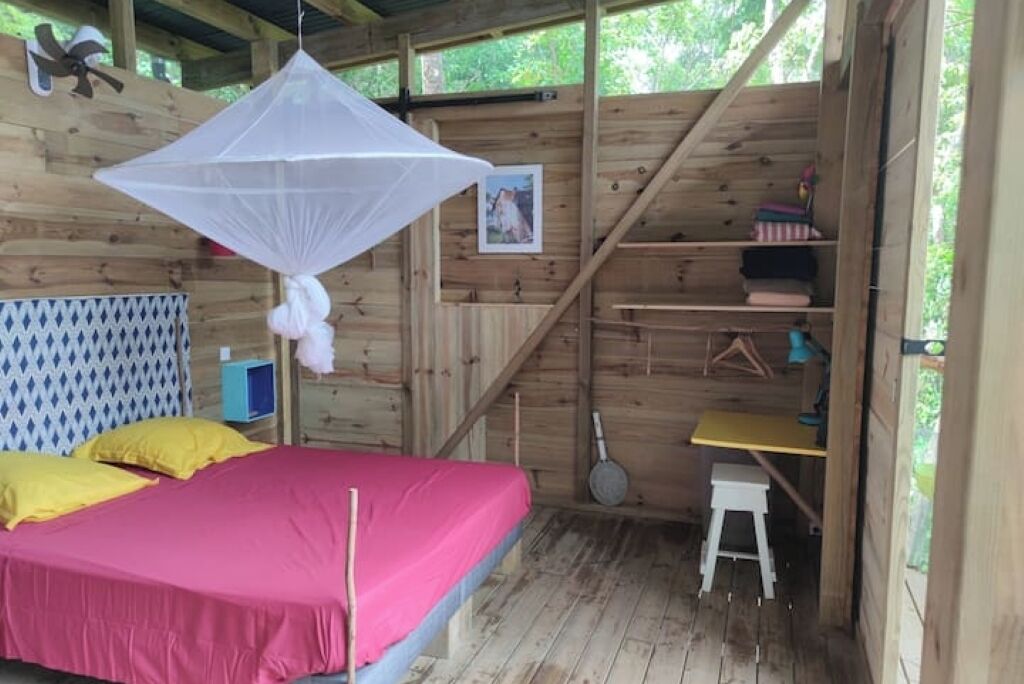 Eine Holzhütte mit einem Bett, einem hängenden Moskitonetz, einem kleinen Hocker und einem Regal mit gefalteter Kleidung.