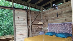 Une cabane en bois avec un lit, une étagère où sont suspendus des vêtements et un escalier.