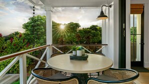 A round table with wicker chairs and a potted plant on a balcony with a view of lush greenery and a sunset.