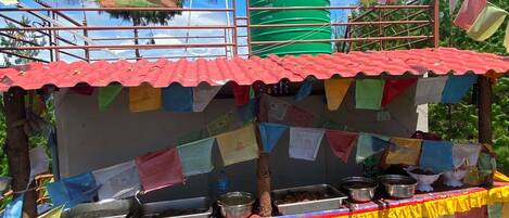 A food stall with a red roof, various cooking utensils, and colorful prayer flags.
