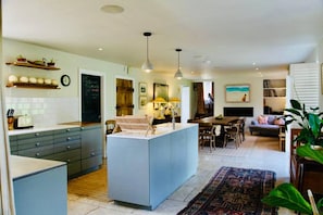 A modern kitchen with a central island, a dining area with a table and chairs, and a living room with a sofa and a large plant. - Cotswold Country House in Charlbury (Charlbury)