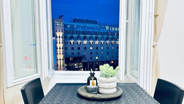 A dining area with a table and chairs, a window with a view of a building labeled "WIMBERT," and a small potted plant on the table. - Elegant 4BR Apt Near Westbahnhof (Vienna)
