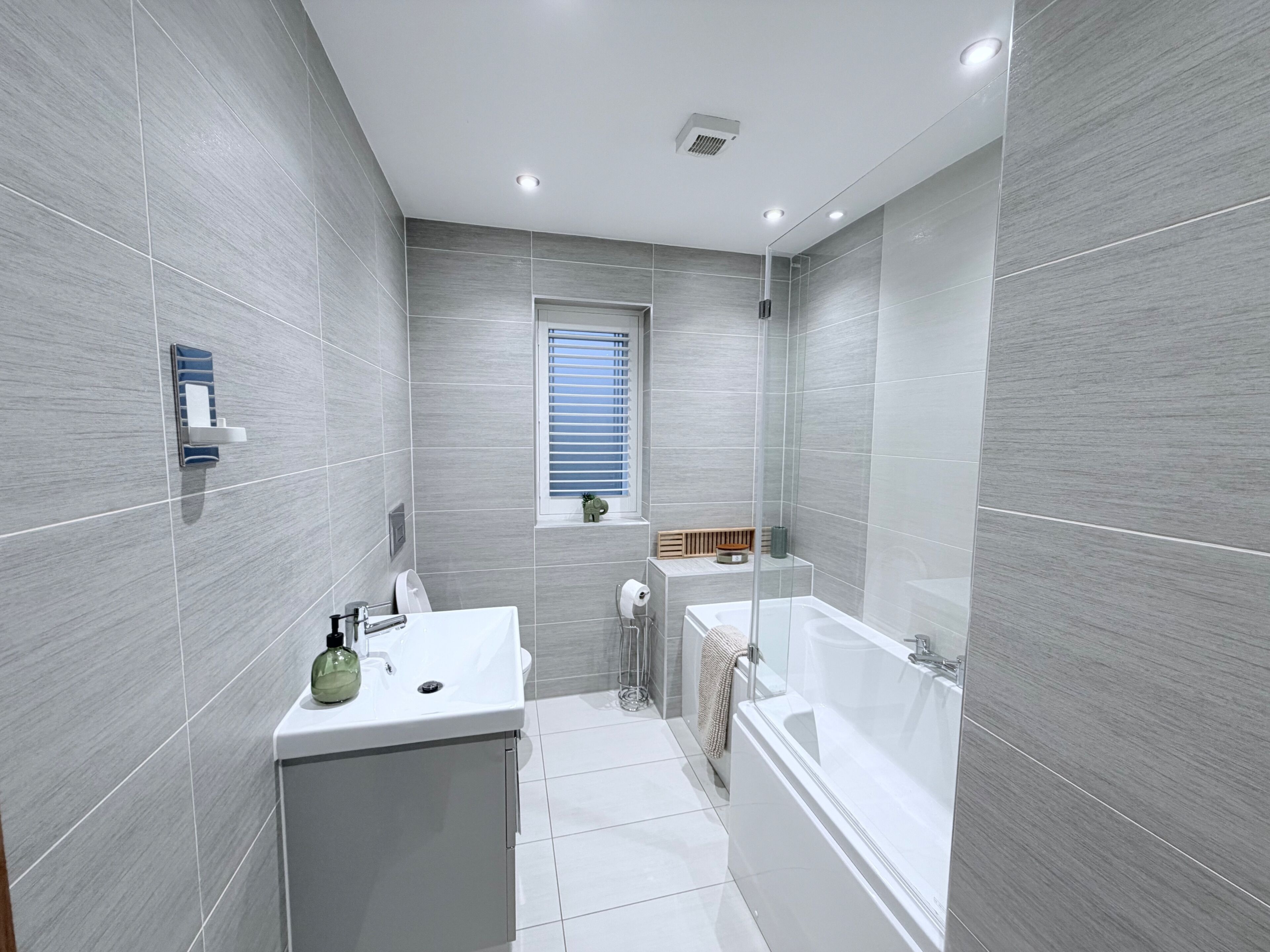 Een moderne badkamer met grijze tegels, een witte wastafel, een douche met glazen wanden en een raam met zonwering.