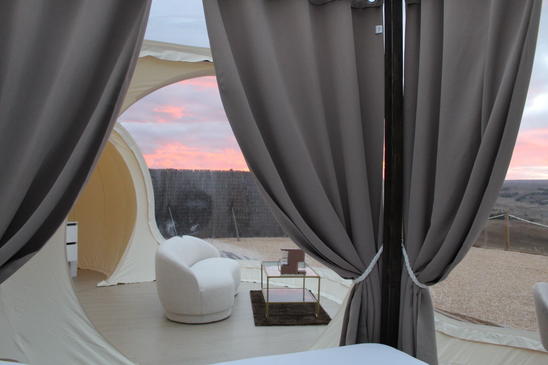 A tented room with a white chair and a small table, a view of a forested landscape, and a sunset.