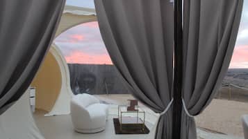 A tented room with a white chair and a small table, a view of a forested landscape, and a sunset.
