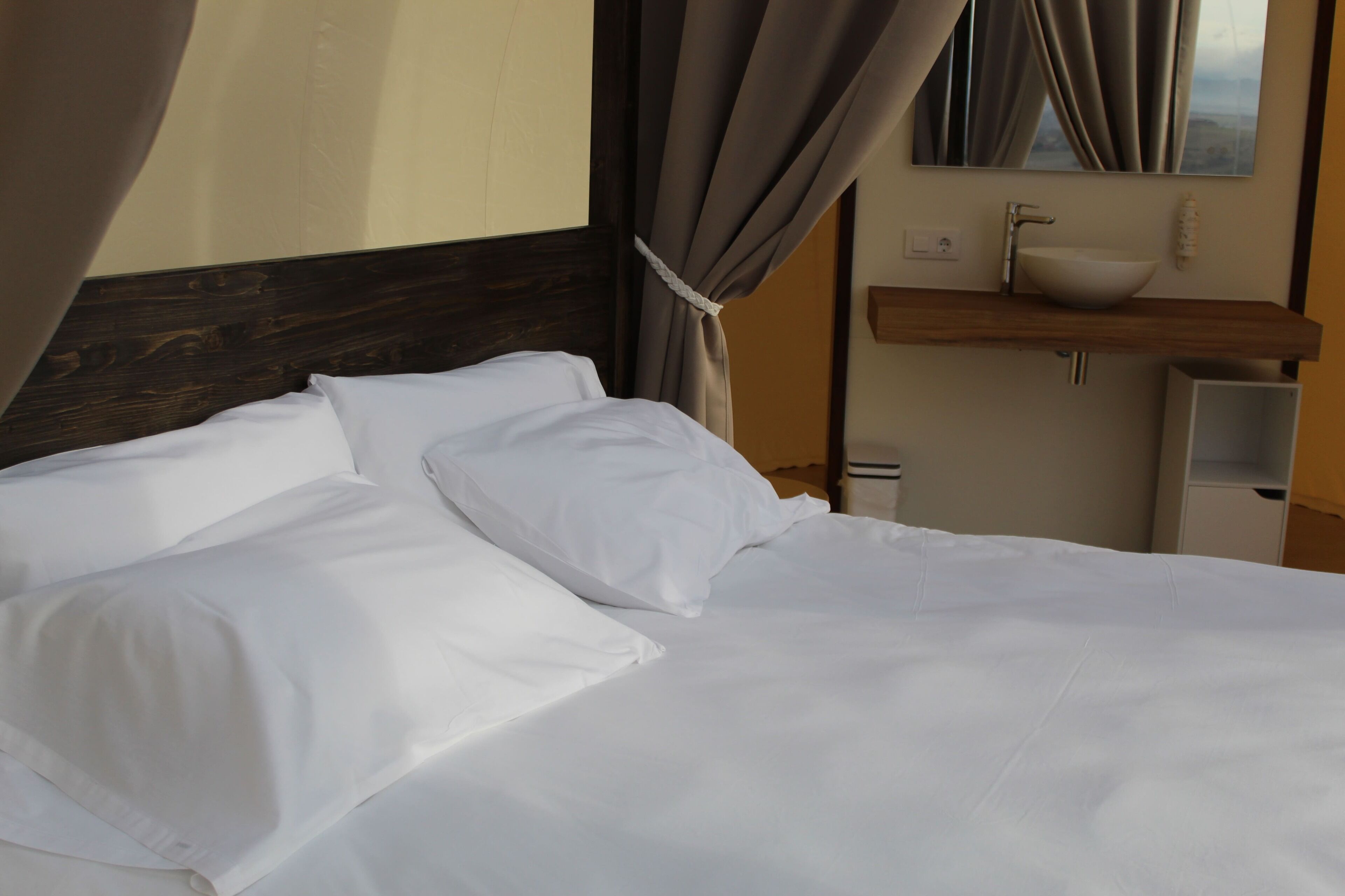 Una cama bien hecha con sábanas blancas, cabecera de madera, mesita de noche con lavamanos blanco y espejo encima.