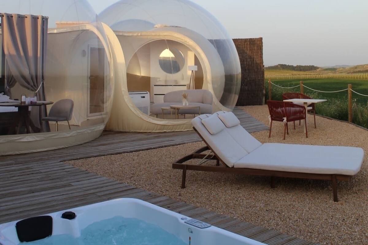 A transparent dome structure with a lounge area, a hot tub, and a view of the countryside.
