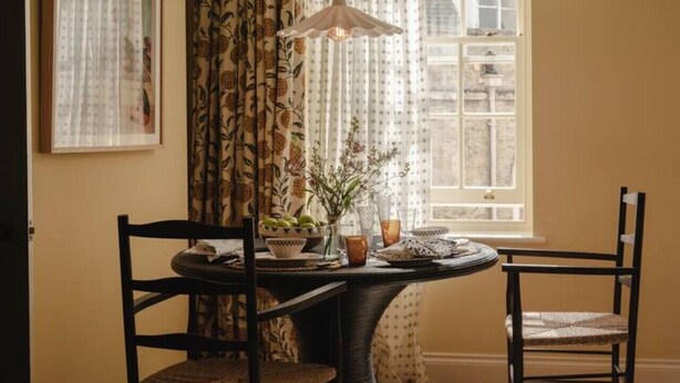 A dining area with a round table, two chairs, a hanging light fixture, and a window with curtains. - Luxury Stay in Heart of the Cotswolds (Cheltenham)