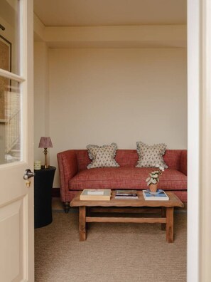 Ein gemütliches Wohnzimmer mit einem roten Sofa, gemusterten Kissen, einem Holztisch mit Büchern und einer Topfpflanze sowie einer Lampe auf einem Beistelltisch.