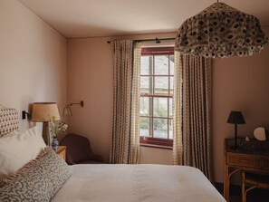 Ein Schlafzimmer mit einem großen Bett, einem Fenster mit Vorhängen, einem Schreibtisch mit Lampe und einem Sessel.