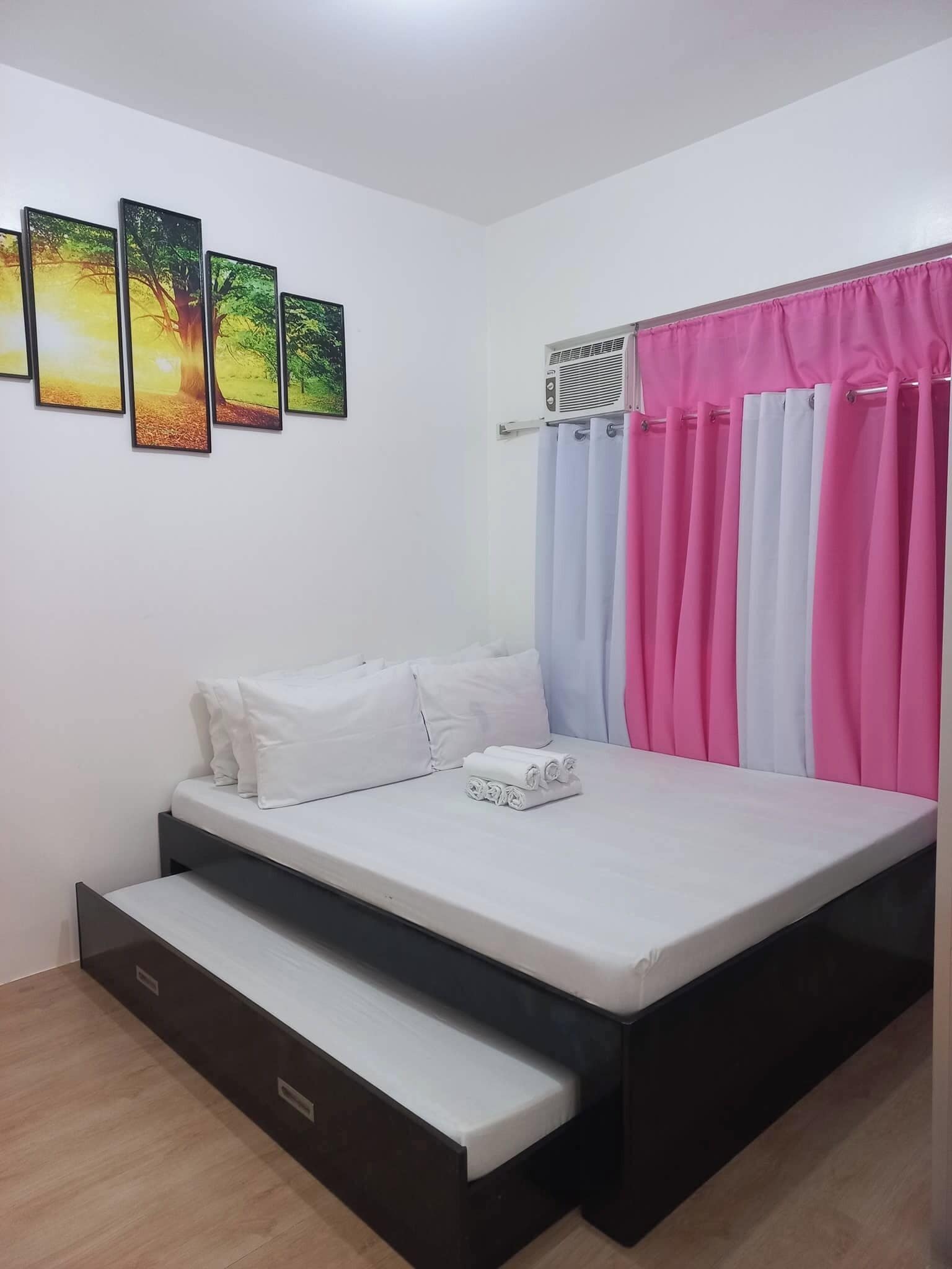 Une chambre à coucher avec un lit, une fenêtre ornée de rideaux roses, un climatiseur fixé au mur et une série de tableaux encadrés.