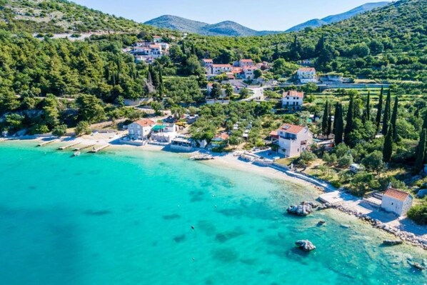 A coastal village with houses along the shoreline, clear turquoise waters, and a hilly backdrop.