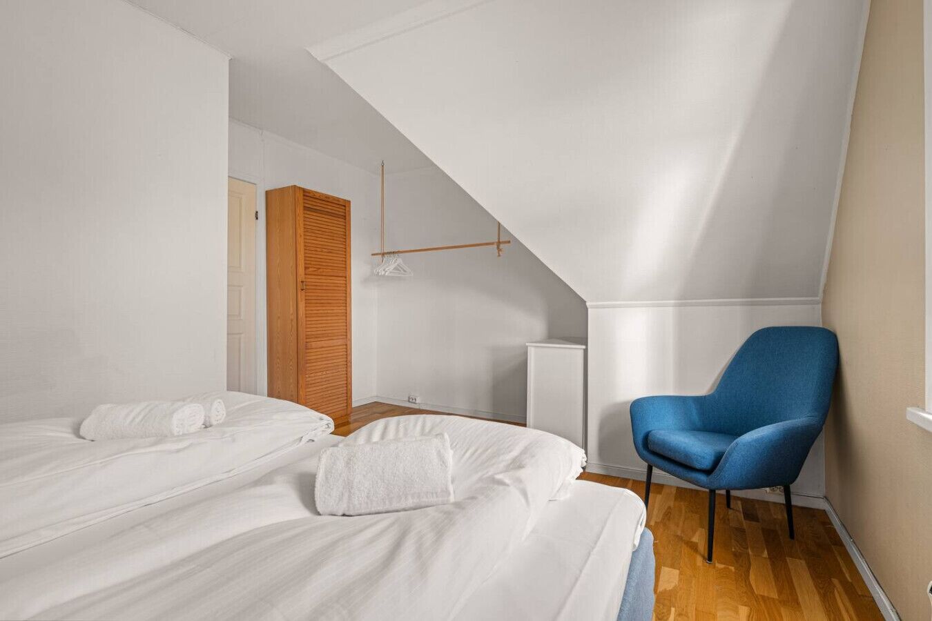 A bedroom with two beds, a blue chair, and a wooden wardrobe.
