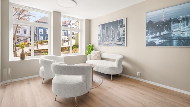 A modern living room with a sofa and armchairs, large windows with a view of buildings, and two cityscape paintings on the wall. - Dream Apartment – Central Living at Its Best! (Bergen)