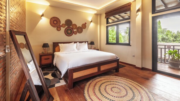 A bedroom with a large bed, a wooden floor, a patterned rug, a wall with circular decorations, and a window with a view of greenery. - 3BR Boutique Villa, Private Pool (Krong Siem Reap)