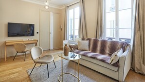 Ein Wohnzimmer mit einer Couch, zwei Sesseln, einem Couchtisch, einem Fernseher und einem großen Fenster mit Vorhängen.