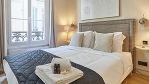 Ein Hotelzimmer mit einem großen Bett, einem Fenster mit Blick auf Gebäude, einem Nachttisch mit Lampe und einem an der Wand hängenden Kunstwerk.
