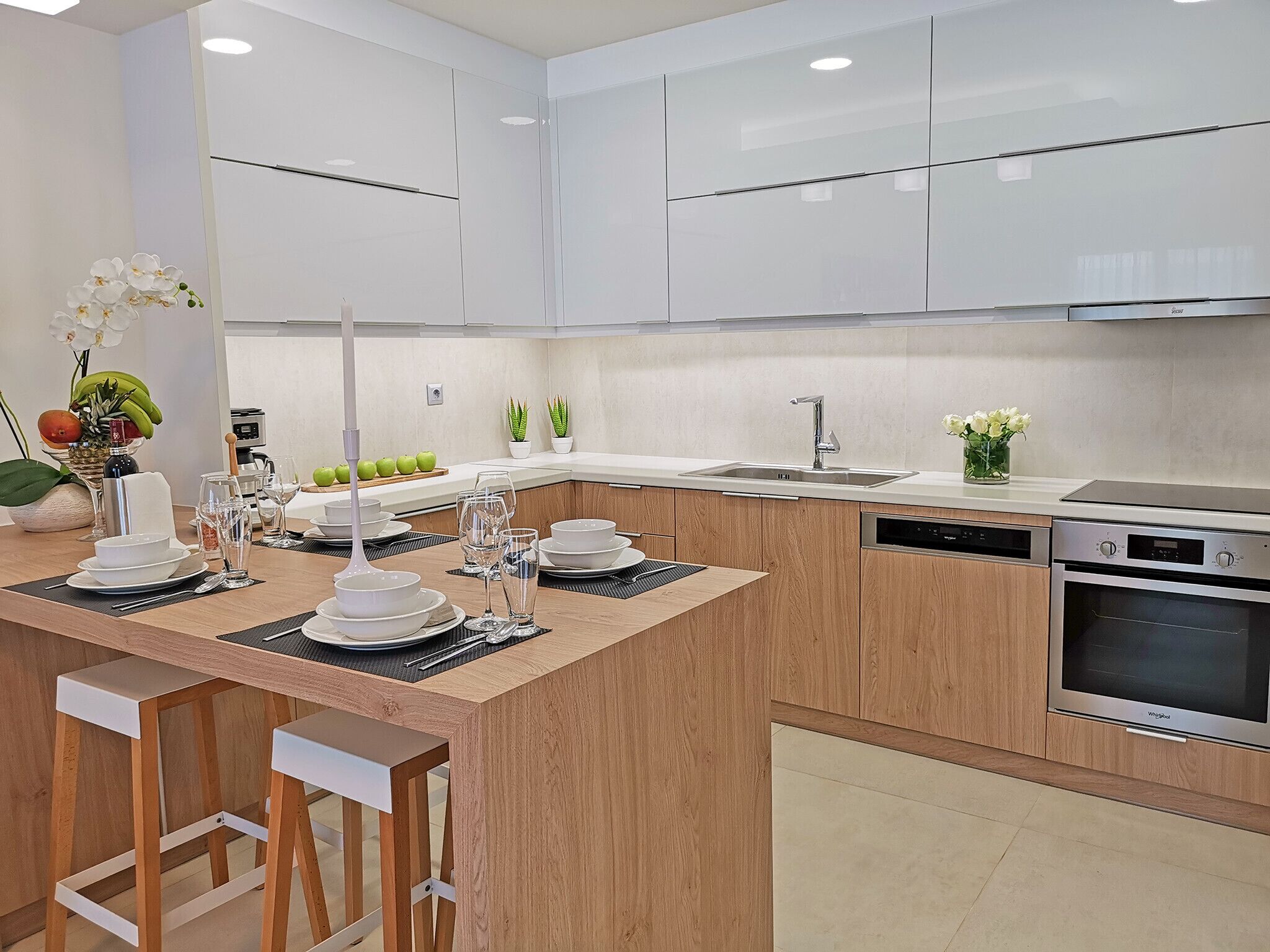 A modern kitchen with white cabinets, a central island with stools, a built-in oven, and a dishwasher.