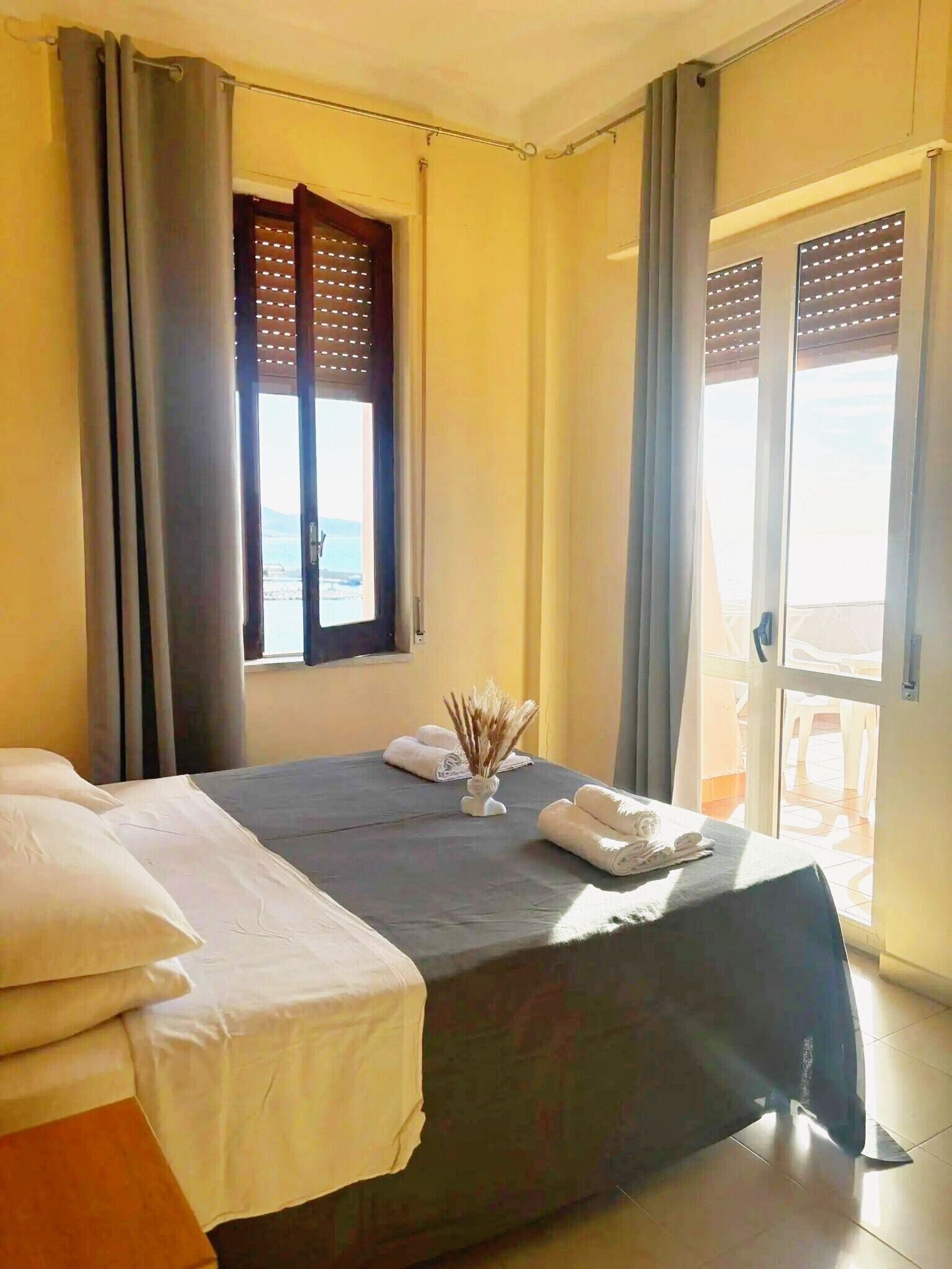 Et hotelværelse med en seng, et vindue med skodder, en dør der fører ud til en balkon, og en lille vase med tørrede planter på sengen.