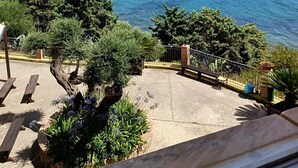 Vista costera con cielo despejado, un muelle que se adentra en el mar y un patio con bancos y plantas en macetas.