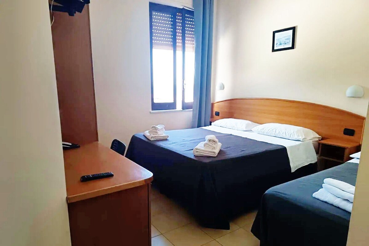 Een hotelkamer met twee bedden, een bureau, een stoel en een raam met jaloezieën.