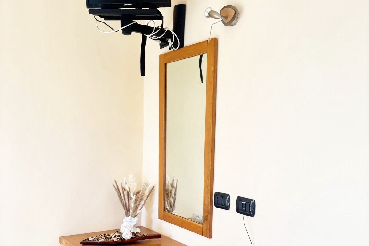 Une petite pièce comprenant un bureau en bois, un miroir, un téléviseur fixé au mur, un téléphone et un petit meuble de rangement.
