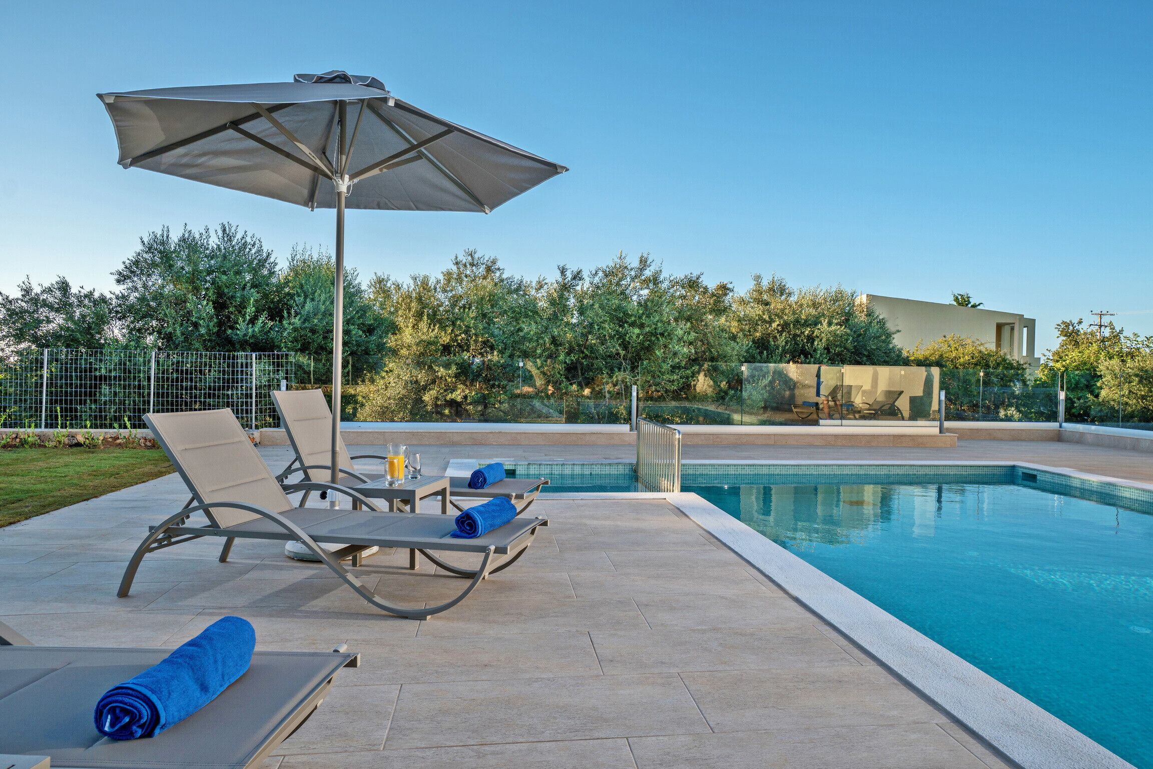 A swimming pool area with lounge chairs and an umbrella, surrounded by a fence and greenery.