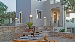 Une table en bois, dressée avec des assiettes, des verres et des bouteilles de vin, est placée sur une terrasse en pierre, avec des marches menant à un bâtiment.