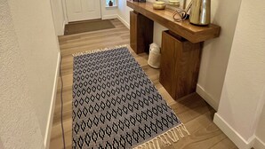 A hallway with a wooden floor, a black and white patterned rug, a wooden console table with a mirror, a vase with green foliage, and a door at the end. - New spacious 4-bedroom house in serene Brighton with cool AC and fast Wi-Fi. (Brighton)