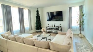 A modern living room with a large beige sectional sofa, a flat-screen TV mounted on the wall, and a small Christmas tree. - New spacious 4-bedroom house in serene Brighton with cool AC and fast Wi-Fi. (Brighton)