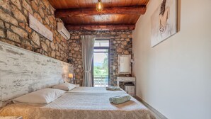 A bedroom with a stone wall, wooden ceiling, a bed with patterned bedding, a nightstand, a desk, and a window with a view. - Souda Bay Cottage (Agios Vasileios)