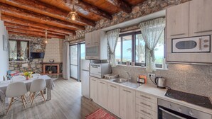 A rustic kitchen with stone walls, wooden ceiling beams, a dining area with a table and chairs, and a built-in oven. - Souda Bay Cottage (Agios Vasileios)