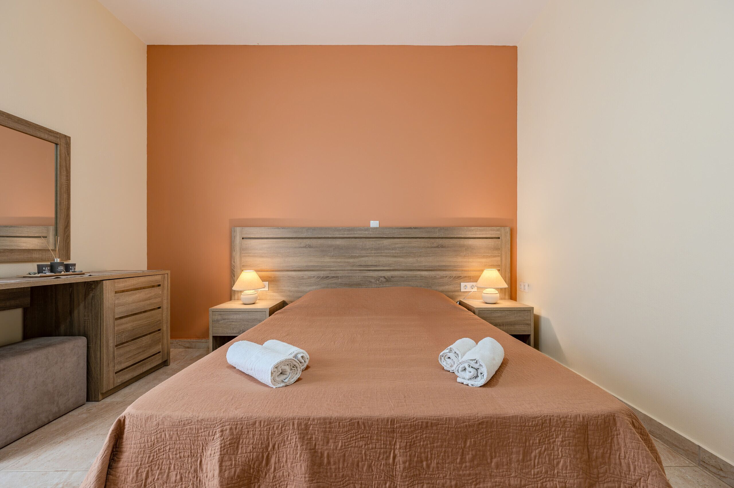 A hotel room with a wooden headboard, two bedside tables with lamps, a bed with a brown bedspread, and a bench at the foot of the bed.