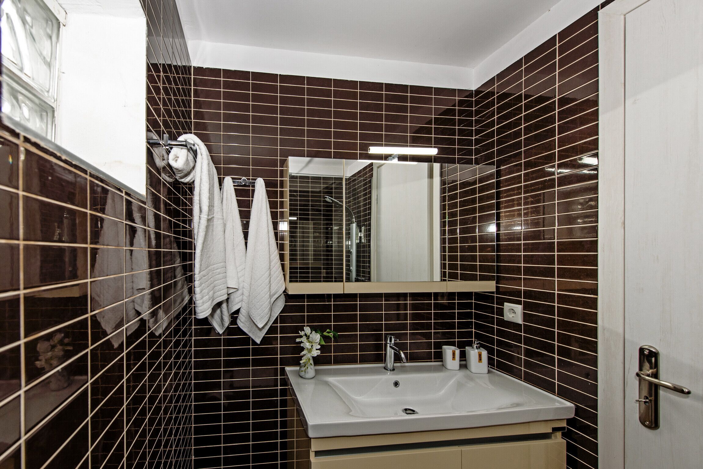 A bathroom with dark brown tiled walls, a white sink, a mirror, and a towel rack with two towels.