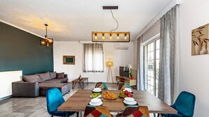 A modern living room with a sofa, dining table, and chairs, a TV, and a window with curtains. - Platanias Villa Spyros (Rethymno)