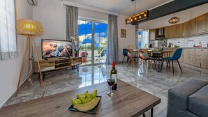 A modern living room with a large TV, a dining area with a table and chairs, and a kitchen with wooden cabinets. - Platanias Villa Spyros (Rethymno)