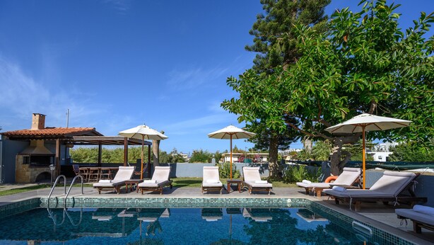 A swimming pool with lounge chairs and umbrellas, surrounded by trees and a clear sky. - Platanias Villa Spyros (Rethymno)