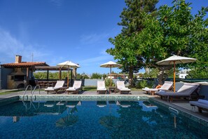 Une piscine entourée d’arbres, avec des chaises longues et des parasols, sous un ciel dégagé.