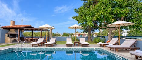 A swimming pool with lounge chairs and umbrellas, surrounded by trees and a building with a tiled roof.
