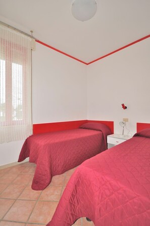Una habitación con dos camas, una ventana con cortinas, una mesita de noche blanca con lámpara y una pared de acento roja.