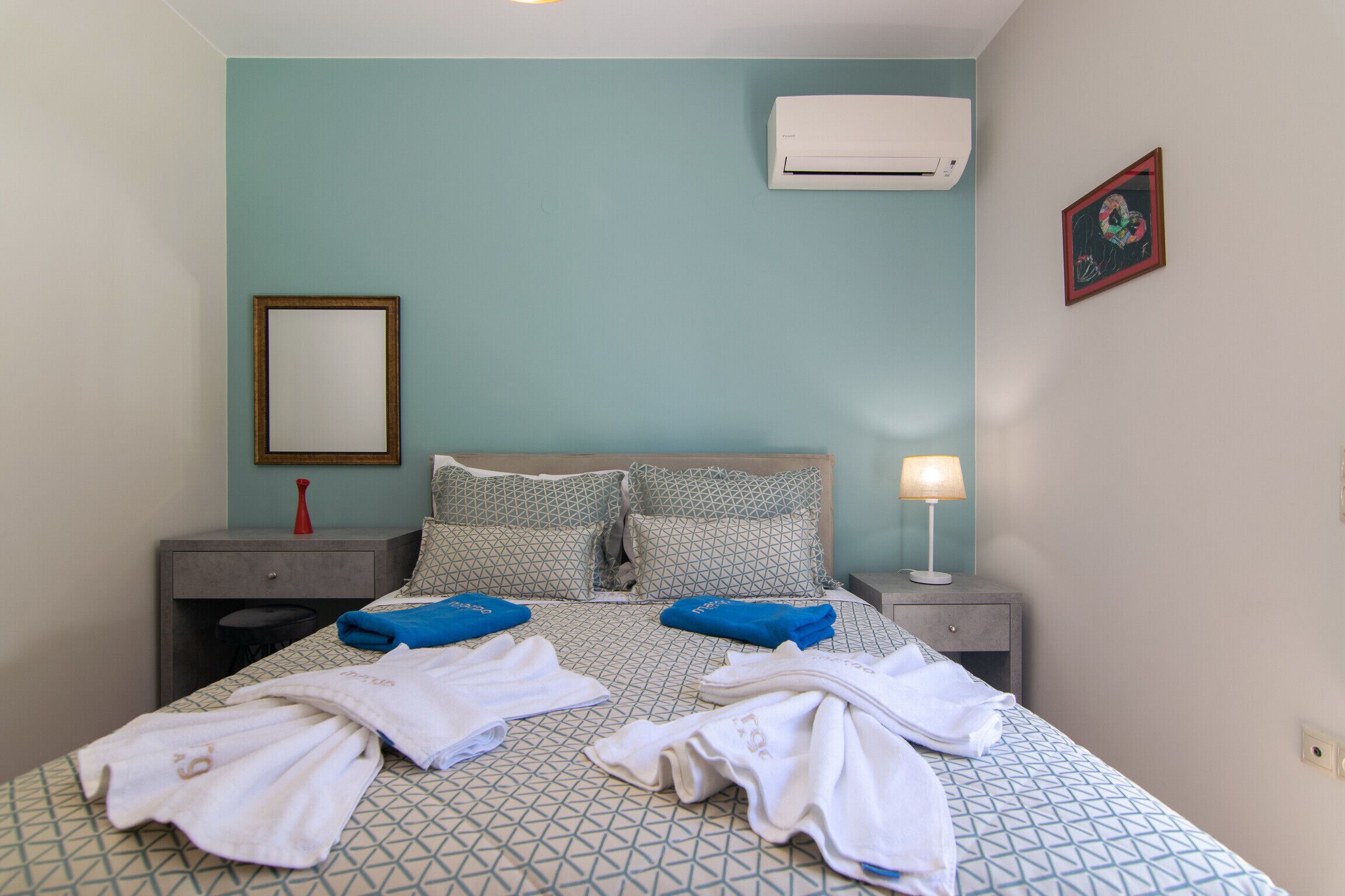 A hotel room with a bed, two nightstands, a bedside lamp, an air conditioner, a framed picture, and a red vase.