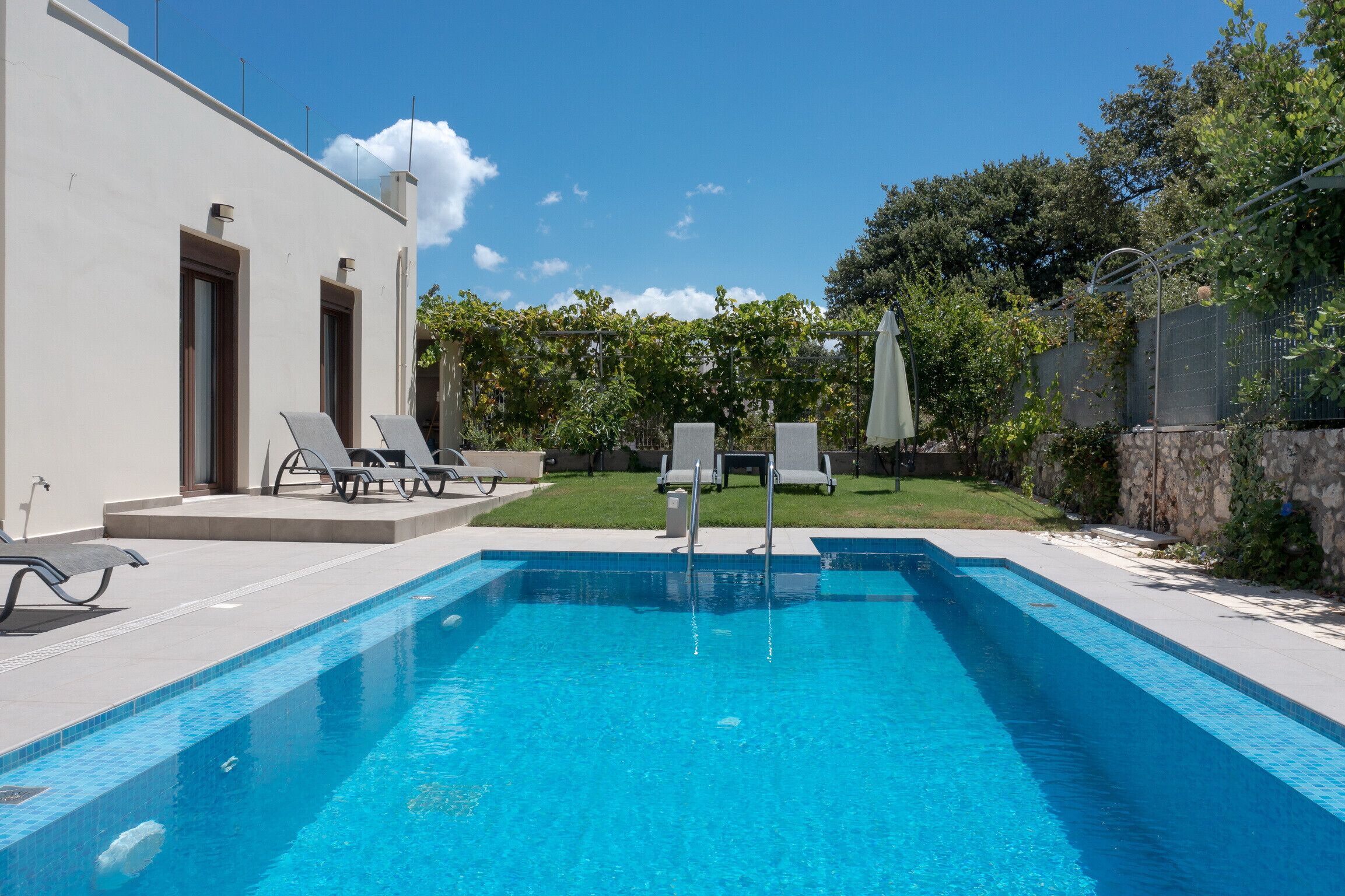 Une piscine entourée d’un mur de pierre et de verdure, avec des chaises longues et des parasols.